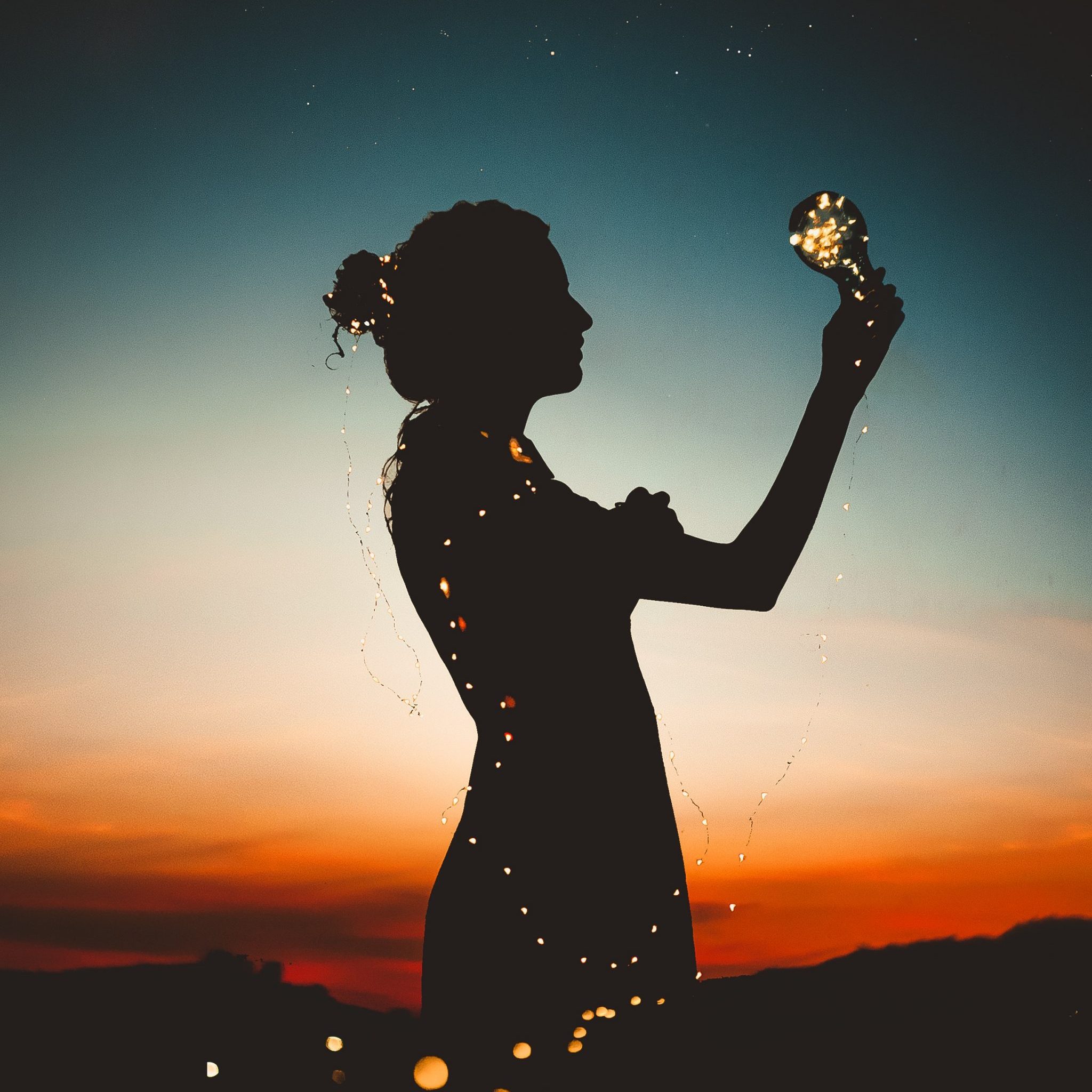 Silhouette holding a glowing bulb wrapped in lights at sunset, symbolizing inspiration Spiritual Awakening